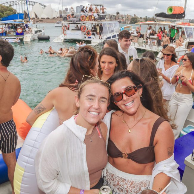 Two smiling women pose for a photo on a boat, surrounded by other people enjoying a lively party on the water with multiple boats, floats, and swimmers in the background.