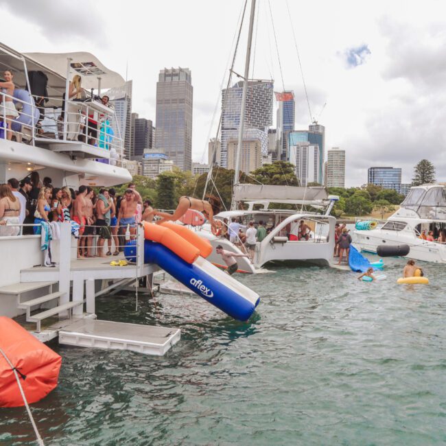 A lively group of people on a docked party boat, some entering the water via inflatable slides, with other boats and city skyscrapers visible in the background on a cloudy day.
