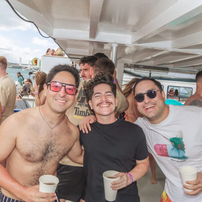 Three men smiling and posing together on a boat party, holding drinks. Other people are mingling and enjoying the event in the background. It appears to be a sunny day on the water.