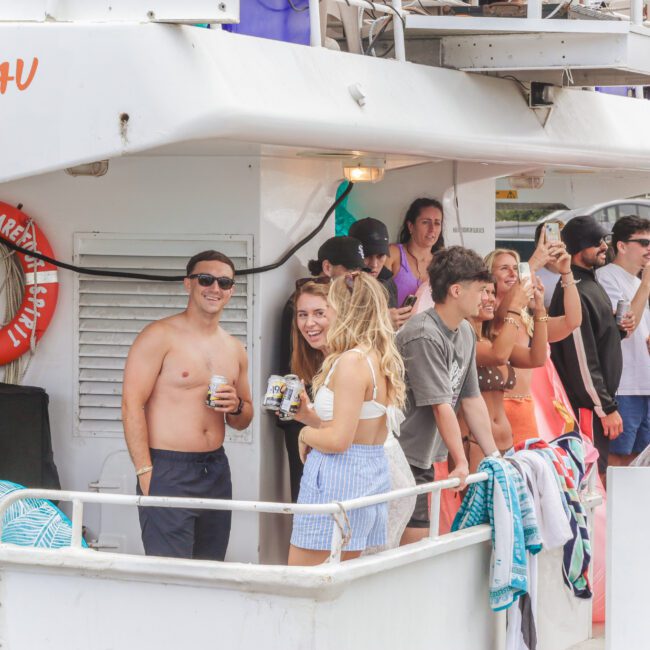 A group of people relax and socialize on the deck of a boat, holding drinks and smiling. Towels hang over the railing, and the weather appears warm and sunny. A life ring and beach items are visible on the boat.