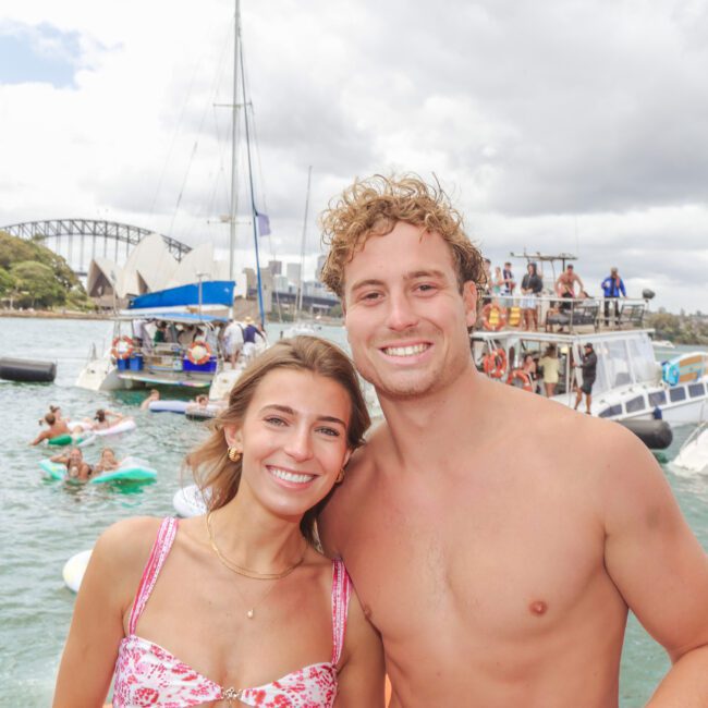 A smiling woman and man pose together at a boat party on the water, with several boats, people, and the Sydney Opera House and Harbour Bridge visible in the background under a cloudy sky.