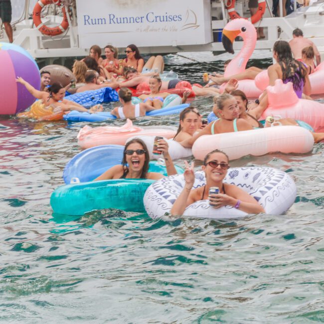 A group of people float on colorful inflatable pool toys in the water near a boat labeled "Rum Runner Cruises," laughing and waving at the camera during a lively summer gathering.