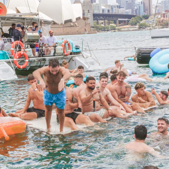 A large group of people in swimsuits enjoy a pool party on the water with inflatables, near boats and the Sydney Opera House in the background on a sunny day.