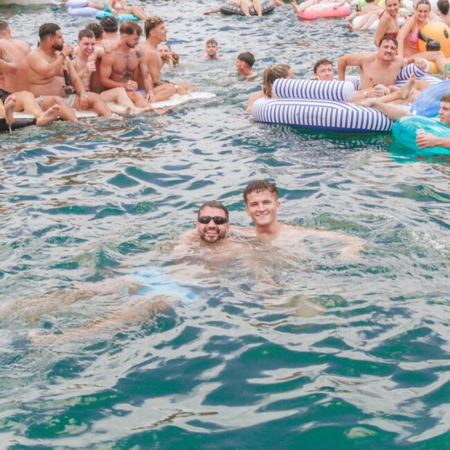 A large group of young adults enjoy a lively pool party, swimming and relaxing on inflatable floats shaped like animals and lounges. Two men smile at the camera while swimming in the foreground.