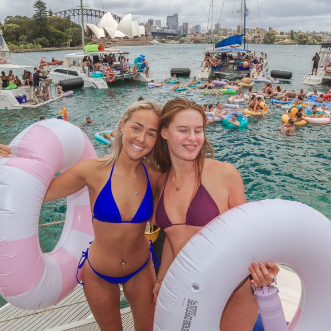 Two women in swimsuits smile and hold inflatable rings on a boat, surrounded by others on floats and boats in a busy harbor with the Sydney Opera House and Harbour Bridge in the background.
