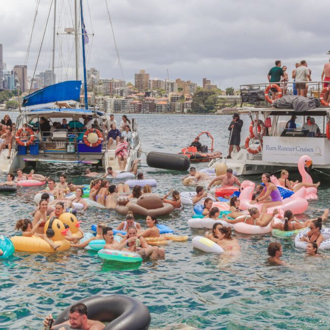 Dozens of people float on colorful inflatable pool toys in a harbor near two docked party boats, with a city skyline and bridge in the background under cloudy skies.