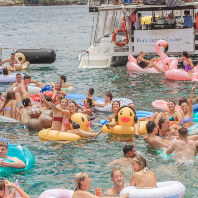 Dozens of people enjoy a sunny day on the water, lounging on colorful inflatable floaties near a boat labeled "Rum Runner Cruises." Some wave and smile, while others relax and swim. The atmosphere is festive and lively.