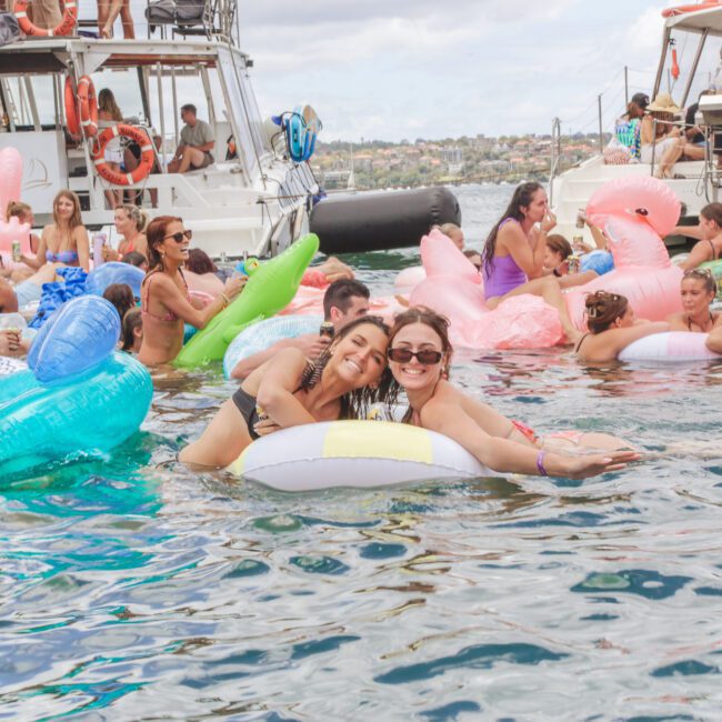 A group of people on colorful inflatable pool floats enjoy a lively party in the water near boats, with many smiling, socializing, and having fun on a sunny day.
