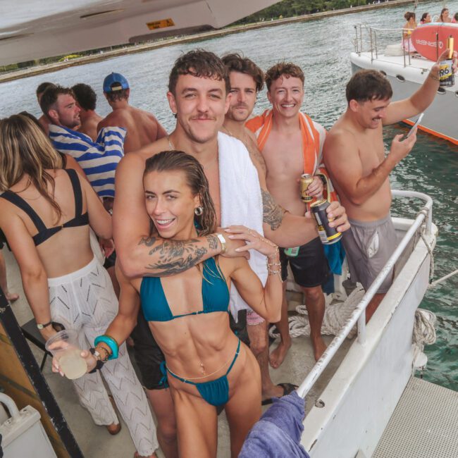 A group of young adults in swimwear smile and pose on a boat, holding drinks and towels. Other people are in the background, some on a nearby boat, and the water and shoreline are visible behind them.