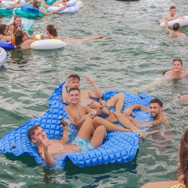 A group of young adults relax on blue inflatable loungers in a crowded body of water, smiling and posing for the camera, surrounded by others on rafts and floaties at a lively pool party.
