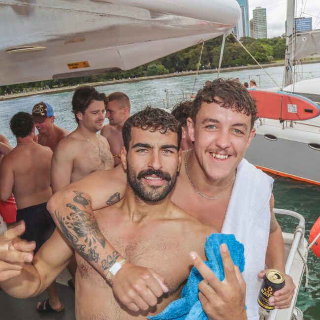 Two smiling men pose on a boat, one with a towel draped over his shoulders, surrounded by a group of people enjoying drinks. In the background, there’s water, another boat, and city buildings.