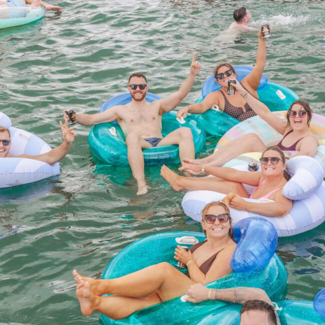 Five people smile and pose with drinks while floating in colorful inflatable tubes on a lake, surrounded by others in the water enjoying a sunny day.