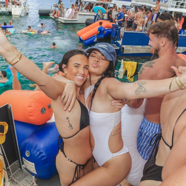 Two women in swimsuits pose happily with arms outstretched on a crowded party boat, surrounded by people swimming and celebrating in the water and on other boats. The atmosphere is lively and festive.