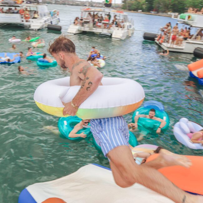 A man wearing striped swim trunks and an inflatable ring jumps into a lake at a lively boat party with people on floats and multiple yachts in the background.