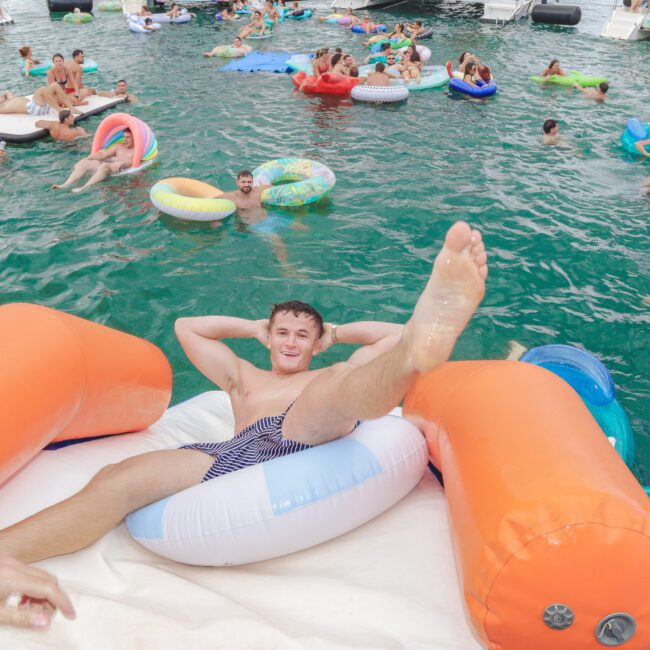 A man relaxes and poses on an inflatable raft with his leg up, smiling at the camera, surrounded by people floating on pool toys in a lively, crowded water scene near boats.