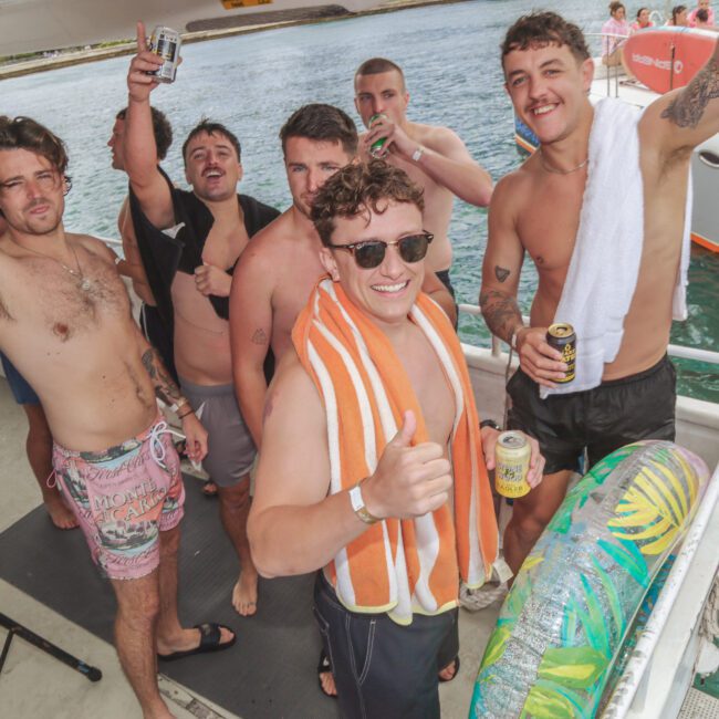 A group of smiling men in swim trunks pose on a boat, holding drinks and giving thumbs up. One has a towel over his shoulders, and another has a towel around his neck. Water and another boat are visible in the background.