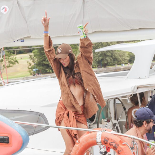 A woman in a brown outfit and cap dances and raises her arms while holding a drink on a boat with other people. A colorful paddleboard and life ring are visible in the foreground. Trees and buildings are in the background.