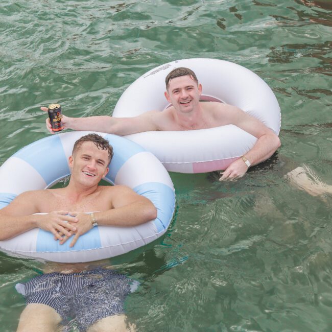 Two men are relaxing in the water on inflatable rings. One man is holding a drink and smiling at the camera, while the other man lounges back, both appearing happy and enjoying a sunny day.