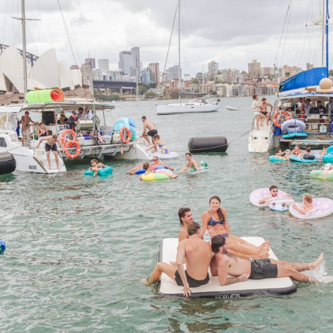 People relax on inflatables and swim in Sydney Harbour near anchored boats, with the Sydney Opera House and Harbour Bridge visible in the background under a cloudy sky.