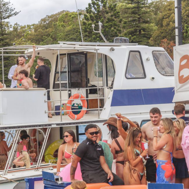 A group of people in swimsuits are gathered on a boat and dock, socializing and enjoying a party. Some are on the boat, while others stand on the dock with drinks. Trees and water are visible in the background.