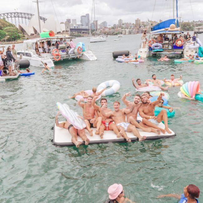 A group of people relax on floating platforms and inflatables in a lively harbor, surrounded by boats. Sydney landmarks, including the Opera House and Harbour Bridge, are visible in the background under a cloudy sky.