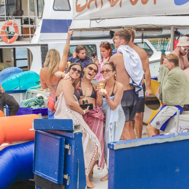 A group of young adults in swimsuits and towels enjoy a lively boat party, smiling and posing for the camera. Inflatable pool floats and drinks are visible, with a yacht and water in the background.