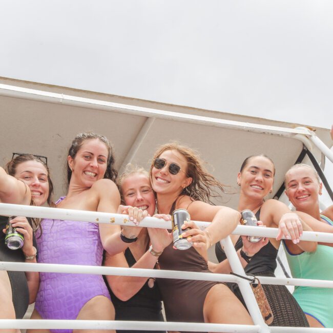 Six women in swimsuits smile and pose together on a boat deck, holding drinks and leaning on the railing, with cloudy skies in the background.