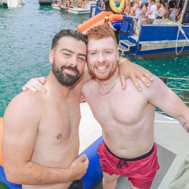 Two smiling men in swim trunks pose arm in arm on a boat deck, surrounded by clear blue water and people enjoying a lively party on boats in the background.