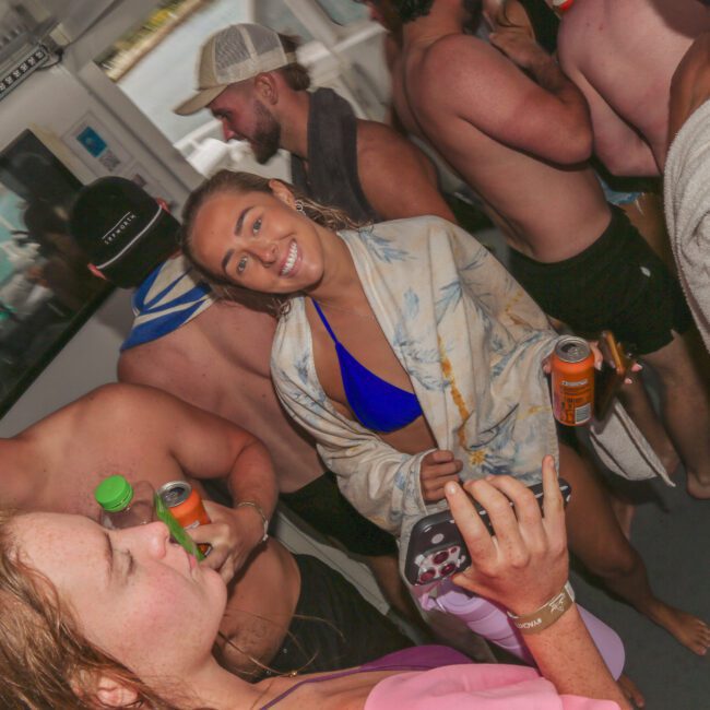 A group of young adults in swimsuits and towels gather closely on a boat, some holding drinks. A woman in the center smiles at the camera while another person in front takes a selfie. The atmosphere is lively and social.