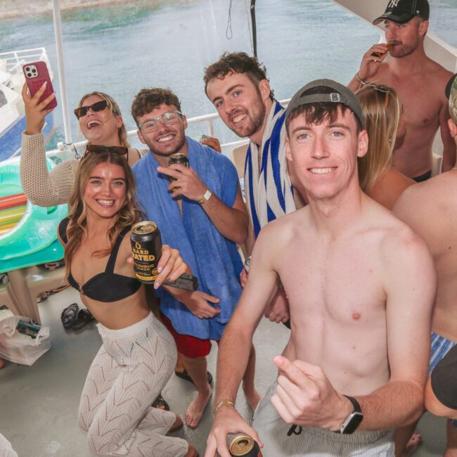 A group of young adults in swimwear smile and pose for a photo on a boat. Some are holding drinks, and the atmosphere is relaxed and festive. Inflatable pool floats and water can be seen in the background.