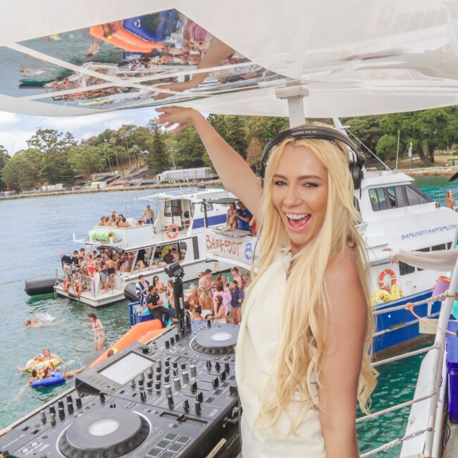 A blonde woman DJ, smiling and wearing headphones, stands at a DJ booth on a boat. Behind her, people are partying on other boats and in the water on inflatables, with trees visible on the shore.