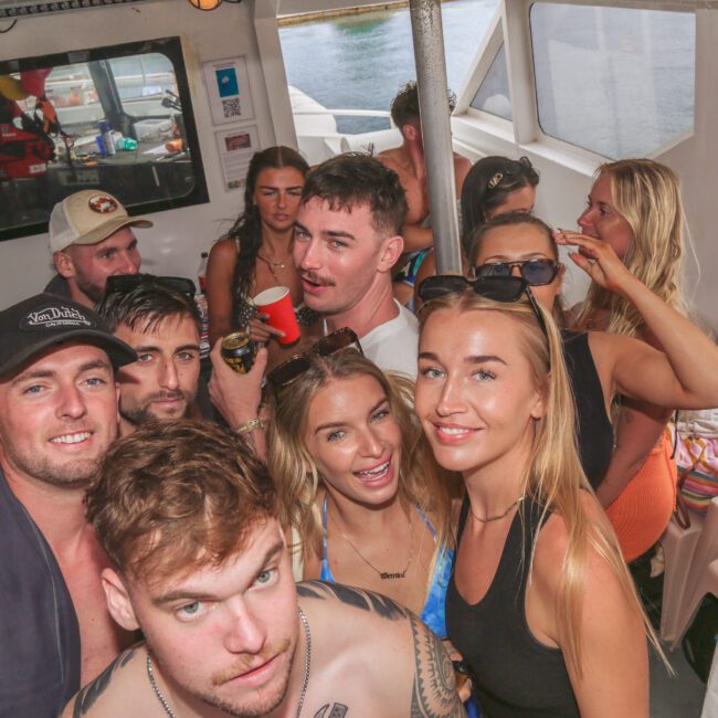 A group of young adults in swimwear smiles and poses for a photo aboard a boat, with water visible through the windows behind them. The mood appears lively and festive.