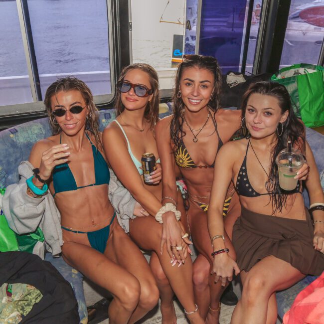 Four women in swimsuits sit closely together on a boat’s cushioned bench, smiling and holding drinks. Bags and personal items are visible around them, and water can be seen through the boat windows in the background.