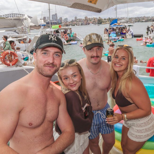 Four young adults, two men and two women in swimwear, smile and pose together on a boat during a lively party. Other boats, people, and inflatables are visible on the water in the background.