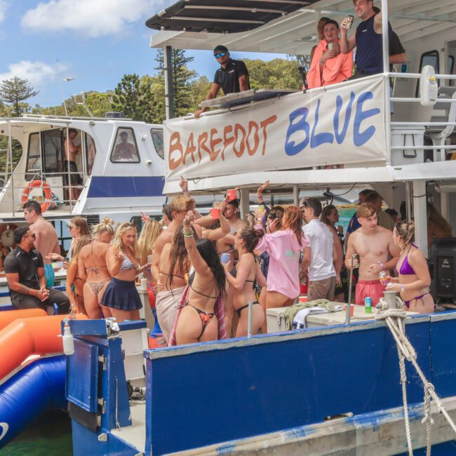 A group of young people in swimsuits are partying on boats named "Barefoot Blue." Some are dancing, others are gathered in groups, and a few are entering the water using an inflatable slide. The scene is lively and festive.