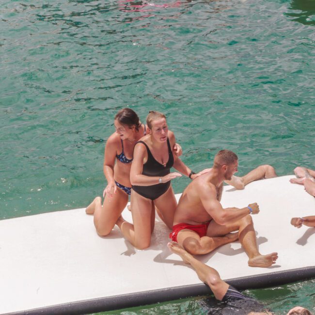 A group of people in swimsuits play and relax on and around a floating white mat in clear green water, some sitting, others laughing, and several swimming nearby. The scene looks lively and fun.