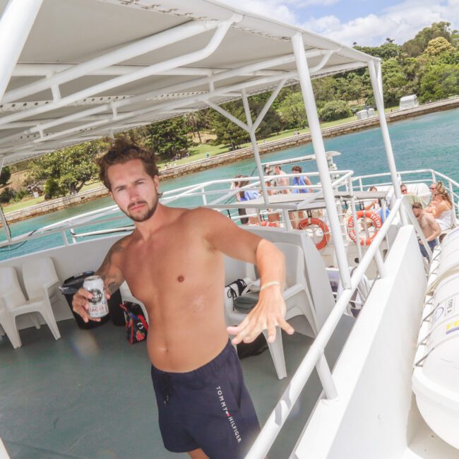 A shirtless man in swim trunks stands on the deck of a boat holding a drink, with clear turquoise water and green trees visible in the background. Other people are seated on the boat's lower deck.