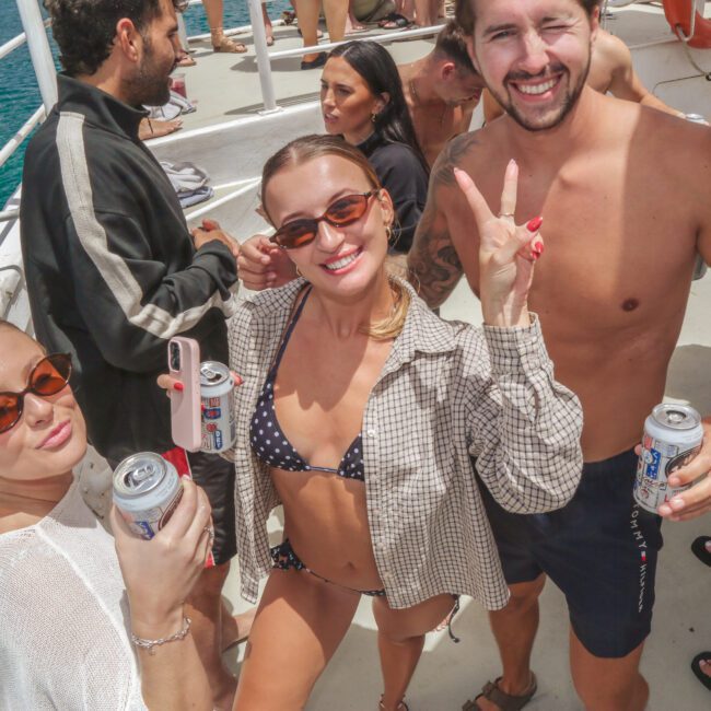 Three smiling people in swimsuits and sunglasses pose with canned drinks on a sunny boat deck, surrounded by others enjoying the water. One woman flashes a peace sign. The scene is lively and bright, with the sea in the background.