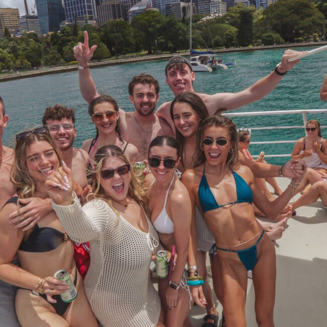 A group of young adults in swimsuits smile and pose for a photo on a boat with drinks in hand. There are buildings and trees in the background across the water, and other people are relaxing on the boat.