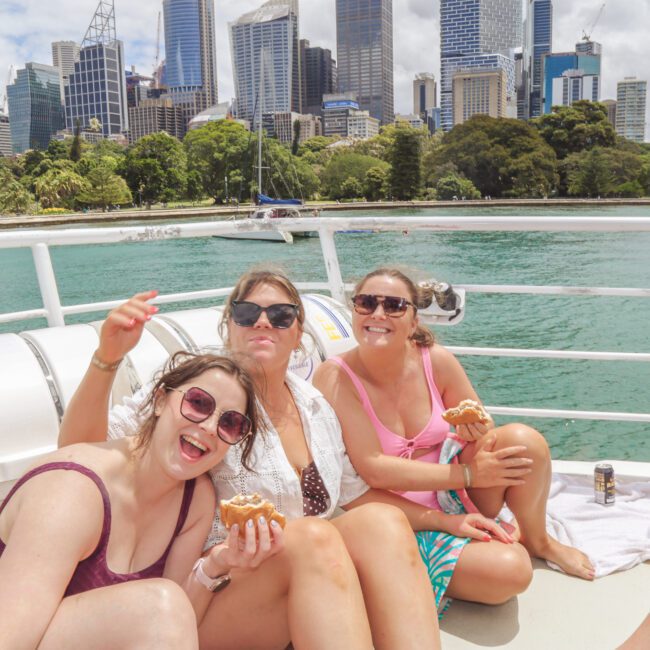 Three women in swimsuits sit on a boat, smiling and eating hot dogs, with a city skyline, green trees, and water in the background on a sunny day.
