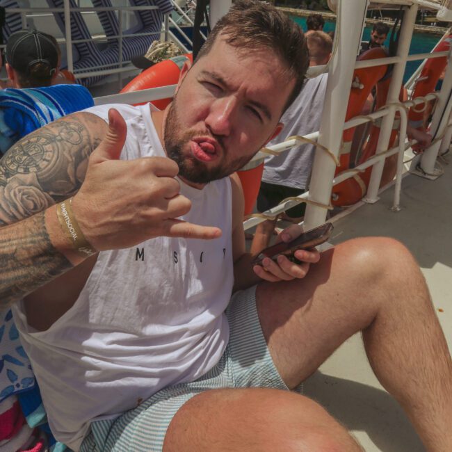 A man in a white tank top and striped shorts sits on a boat deck, making a shaka hand sign and sticking out his tongue. He holds a phone and has drinks beside him. Other people and life vests are visible in the background.