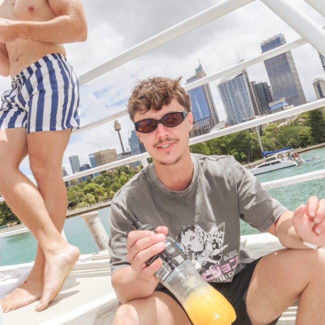 A man wearing sunglasses and a graphic t-shirt sits on a boat holding a drink, with water, city buildings, and another person in striped swim shorts in the background under a partly cloudy sky.