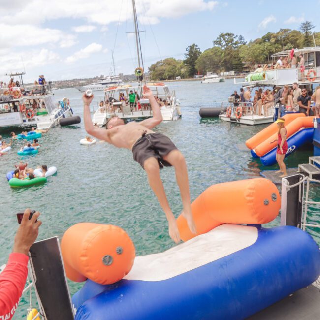 A boy in swim trunks is mid-flip off an inflatable structure into the water, surrounded by boats with people watching and others floating on colorful inflatables, creating a lively summer party scene.