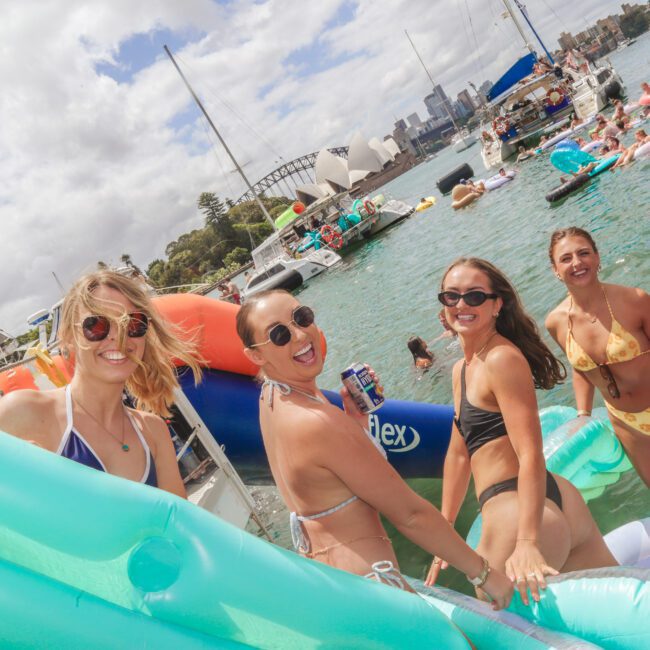 Four women in swimsuits smile at the camera while standing on a pool float in the water, surrounded by other people on inflatables. Boats and the Sydney Harbour Bridge are visible in the background under a partly cloudy sky.