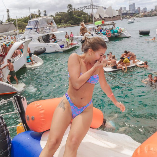 A woman in a bikini laughs while stepping onto an inflatable float at a lively yacht and boat party, with people swimming and relaxing in the water and city skyline visible in the background.
