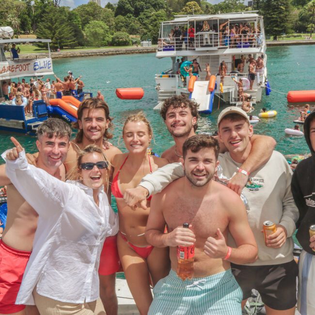 A group of young adults in swimwear smile and pose on a boat, holding drinks. Other people and boats are visible in the water behind them, suggesting a lively summertime party. Trees and greenery are in the background.