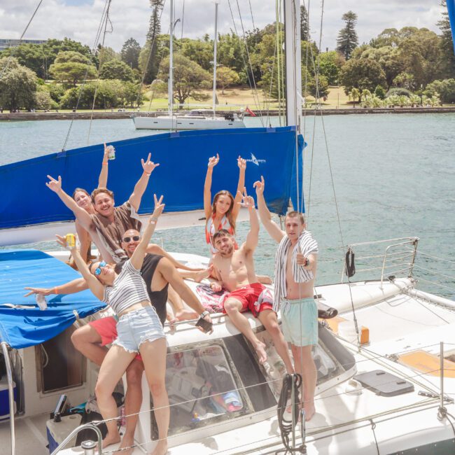 A group of young adults in swimsuits smile and pose with arms raised on the deck of a sailboat, anchored on a sunny day with water, trees, and other boats in the background.