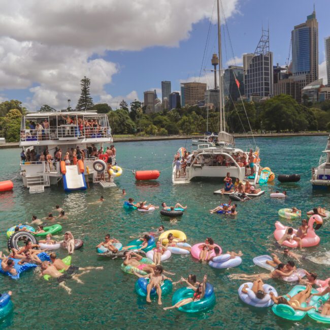 A lively scene of numerous people relaxing on colorful inflatable tubes in turquoise water near boats, with a city skyline and trees in the background under a partly cloudy sky.