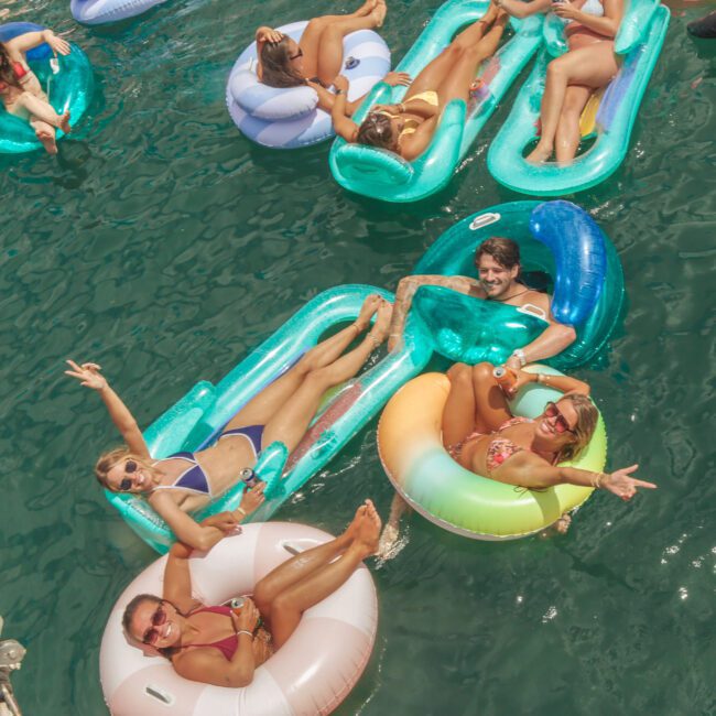 A group of people relax on colorful inflatable floats in clear green water, smiling, waving, and enjoying a sunny day together.