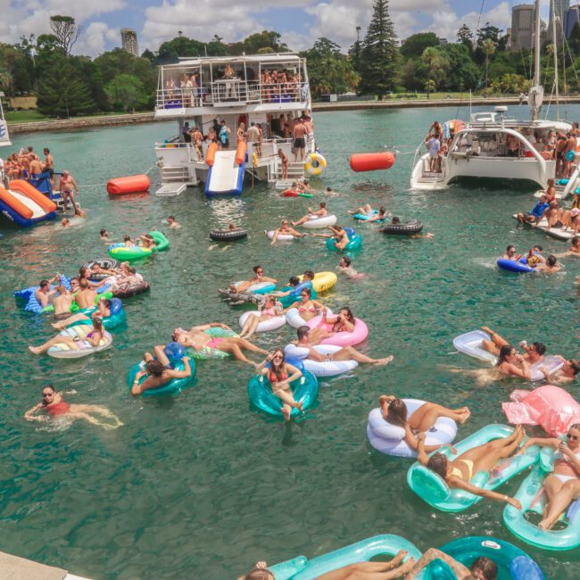 People relax on colorful inflatables in turquoise water between anchored boats, while others gather on deck under a partly cloudy sky with trees and city buildings in the background.